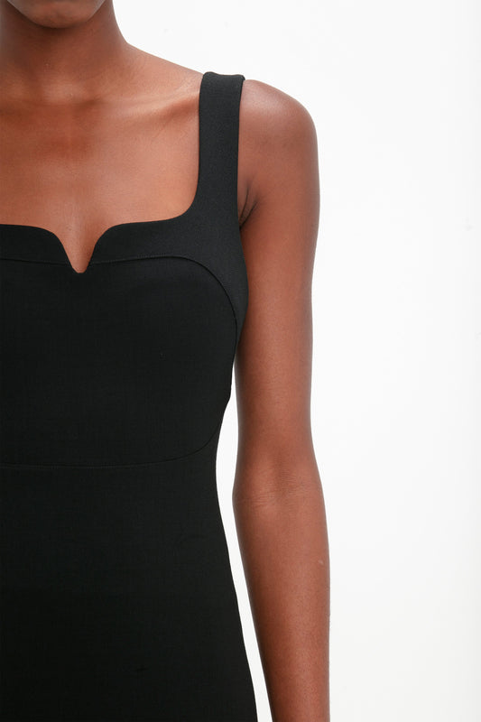 A person wearing a Victoria Beckham Sleeveless Fitted T-Shirt Dress In Black with a square neckline made from stretch double wool crepe. Only their shoulders, chest, and part of the arm are visible against a plain white background.