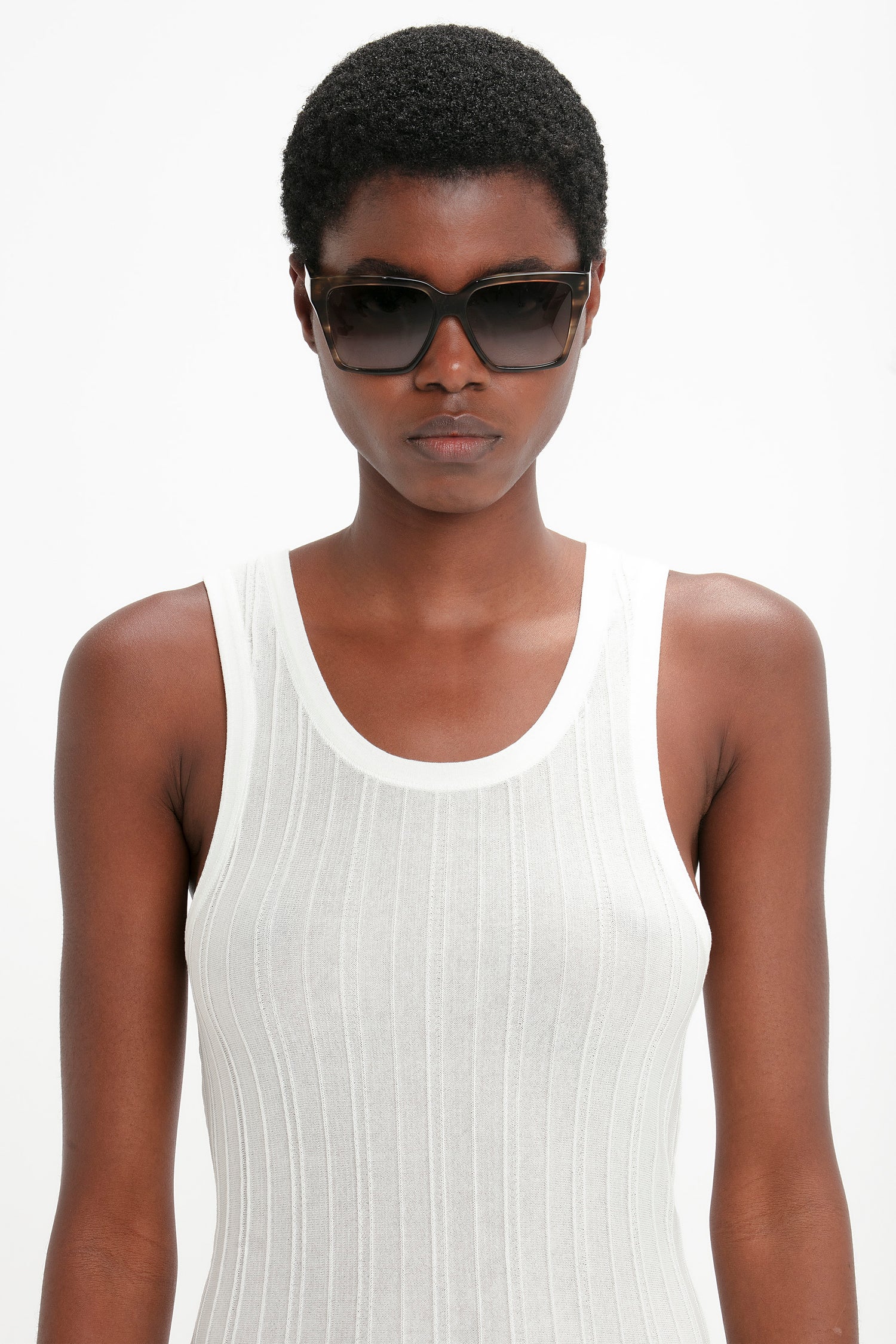 A person wearing Soft Square Frame Sunglasses In Striped Khaki Havana from Victoria Beckham and a white sleeveless top, standing against a plain white background.