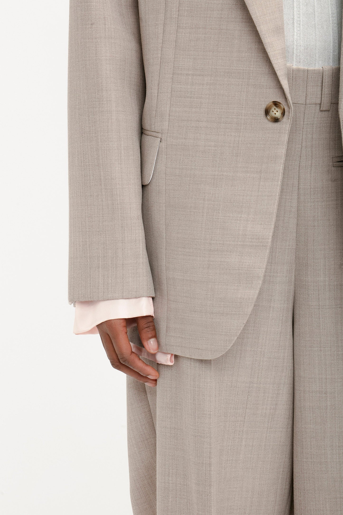 Close-up of a person wearing the Darted Sleeve Tailored Jacket In Sesame from Victoria Beckham with matching trousers. The suit, crafted from pure wool, subtly showcases a light pink shirt sleeve peeking out from under the jacket, highlighting its contemporary detailing.