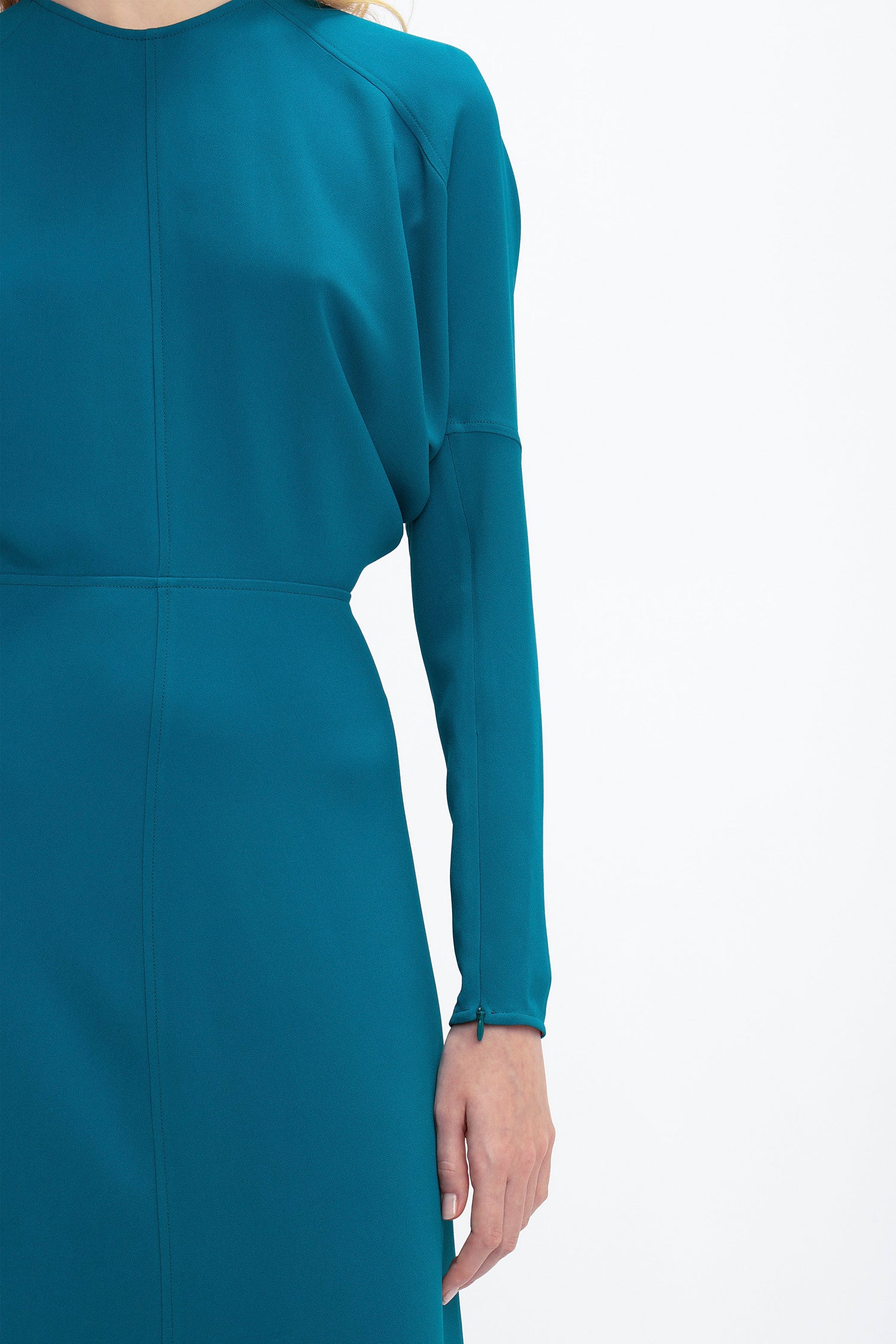 Close-up of a person wearing a Victoria Beckham Long Sleeve Dolman Midi Dress In Petroleum with seam detailing in luxurious cady fabric. Only the upper body and right arm are visible against a plain white background, exuding contemporary sophistication.