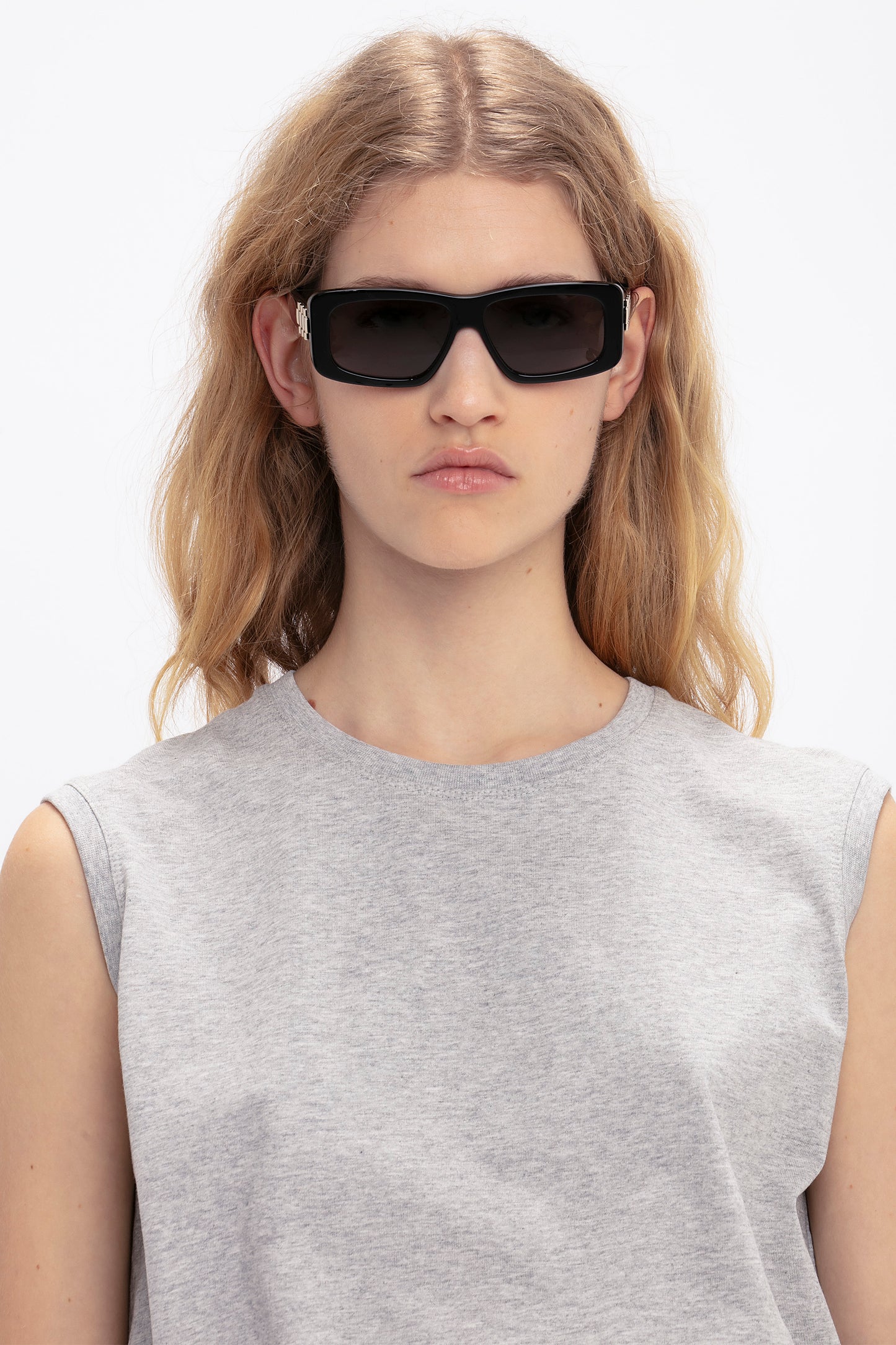 A person with wavy blonde hair wearing Victoria Beckham Chain Detail Rectangular Frame Sunglasses In Black and a light gray sleeveless top against a plain background.