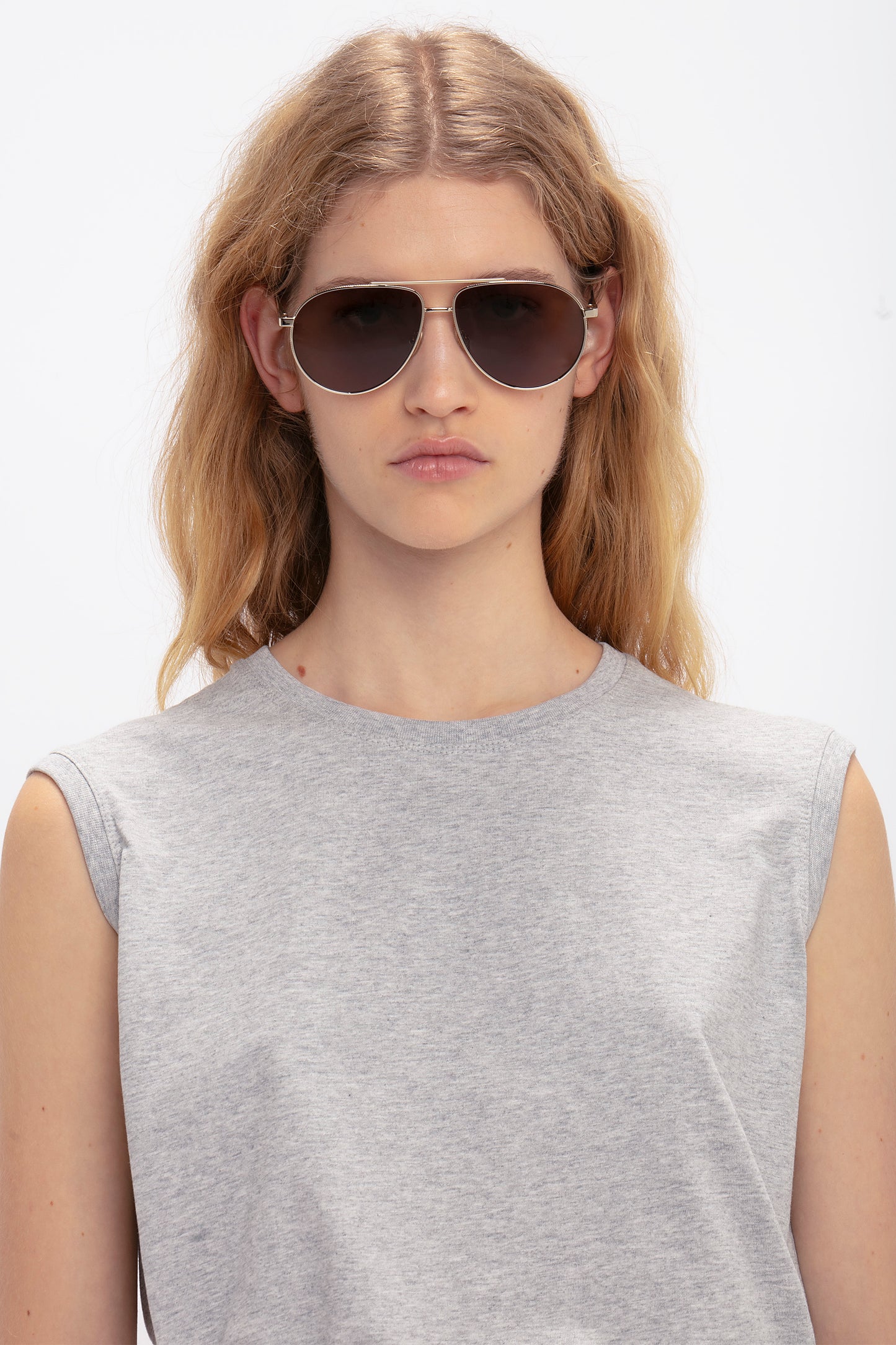 Person with long, loose, blonde hair wearing Victoria Beckham V Metal Pilot Sunglasses In Gold-Khaki with adjustable nose pads and a sleeveless gray top, standing against a plain white background.