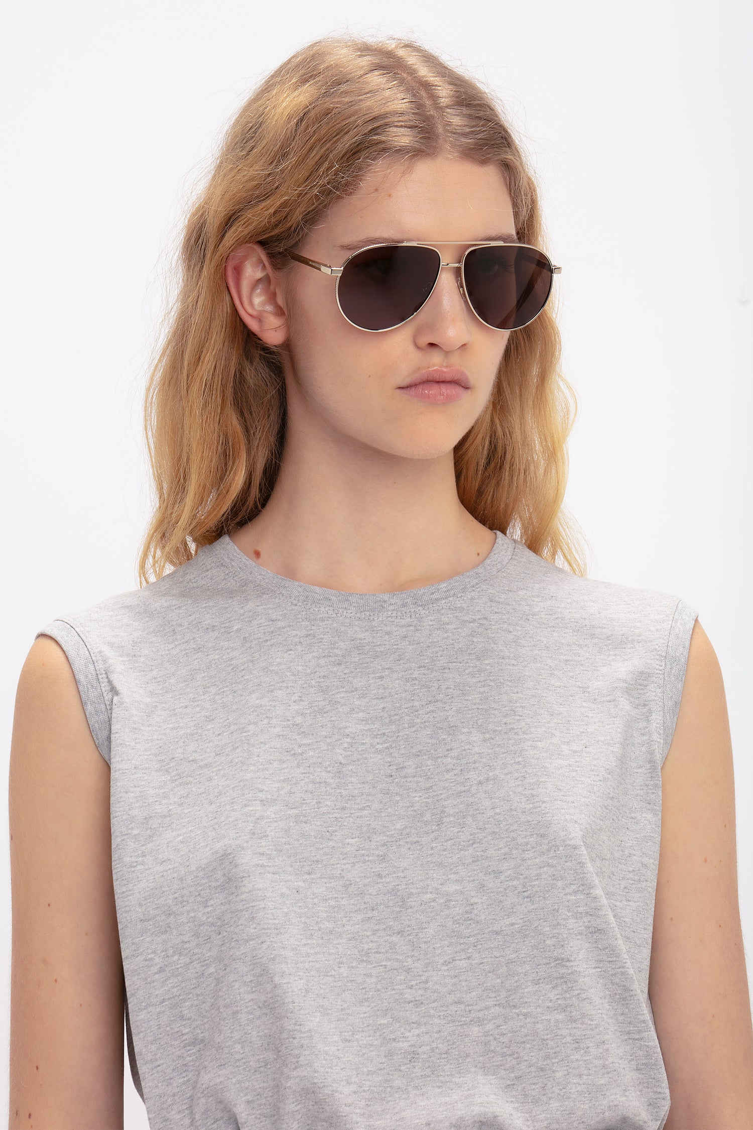 Person with long hair wearing Victoria Beckham V Metal Pilot Sunglasses In Gold-Khaki with adjustable nose pads and a sleeveless gray shirt, looking forward.