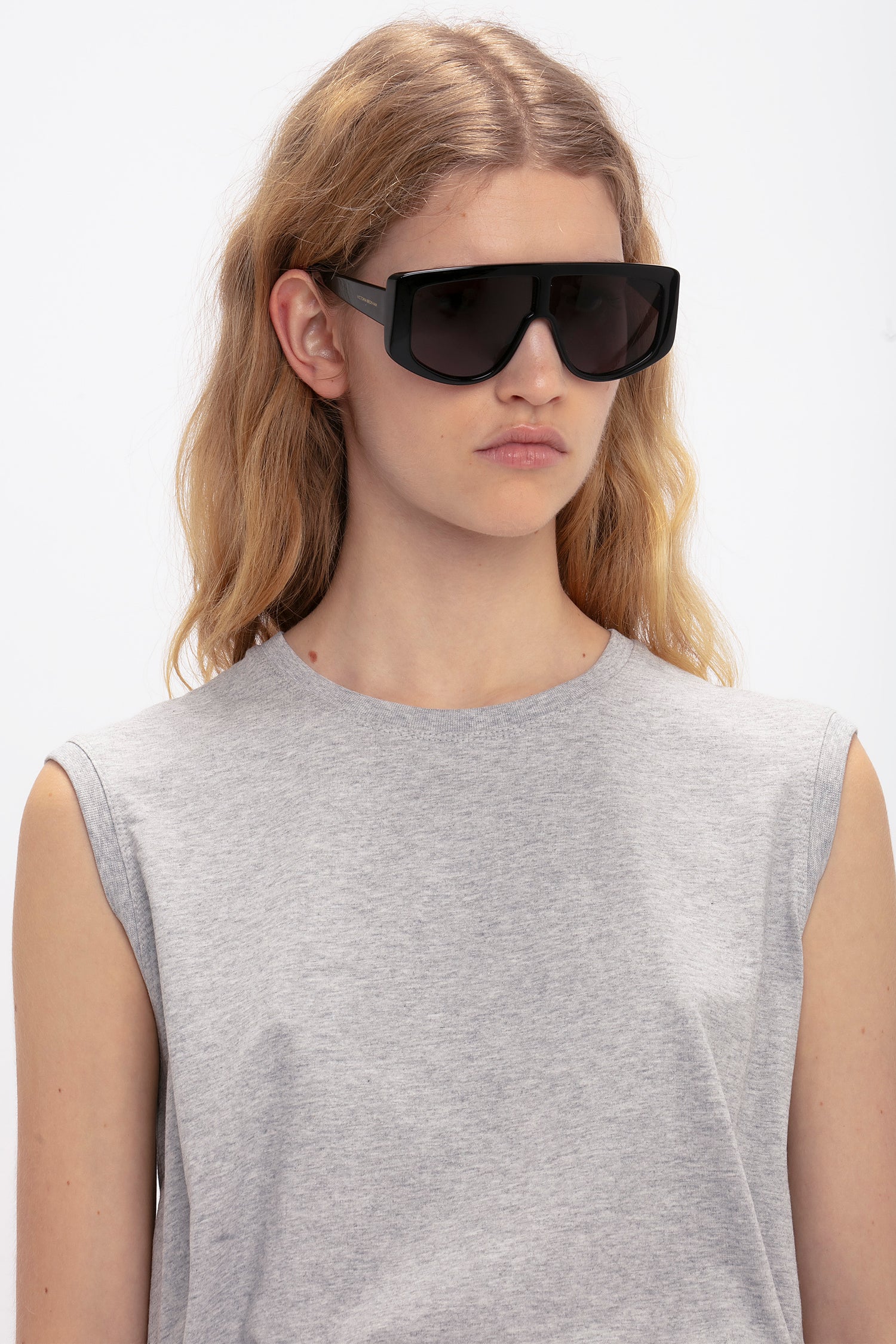 Person with long hair wearing oversized black Acetate Visor Sunglasses In Black by Victoria Beckham and a sleeveless gray top, standing against a plain white background.