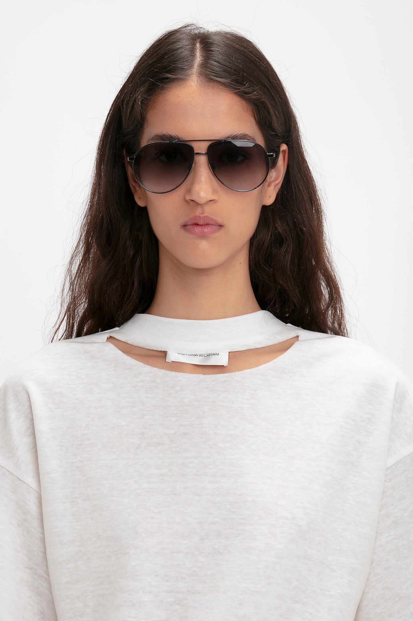 A person with long dark hair, wearing Victoria Beckham V Metal Pilot Sunglasses In Grey Gradient with a gunmetal grey gradient design and a white cutout top, poses against a plain background.