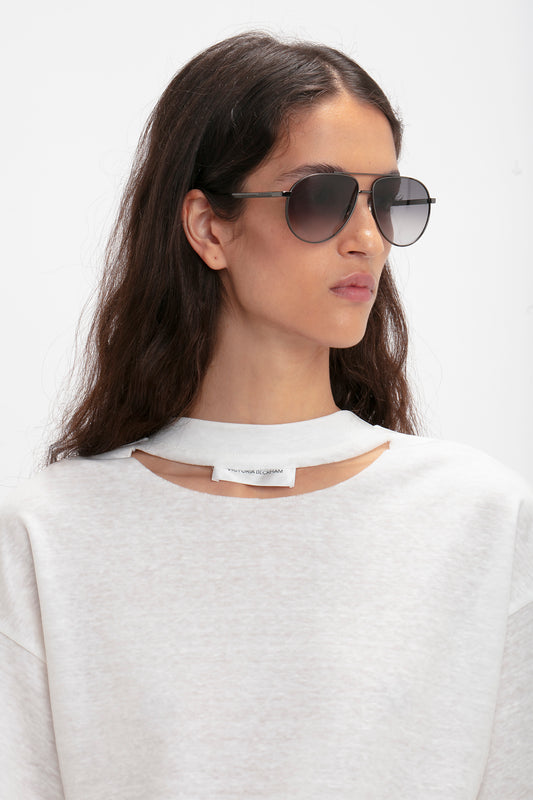 A person with long dark hair wearing Victoria Beckham V Metal Pilot Sunglasses In Grey Gradient and a white top with a small cutout near the collar. The aviators feature a sleek, gunmetal grey gradient design that perfectly complements the look.