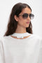 A person with long dark hair wearing Victoria Beckham V Metal Pilot Sunglasses In Grey Gradient and a white top with a small cutout near the collar. The aviators feature a sleek, gunmetal grey gradient design that perfectly complements the look.