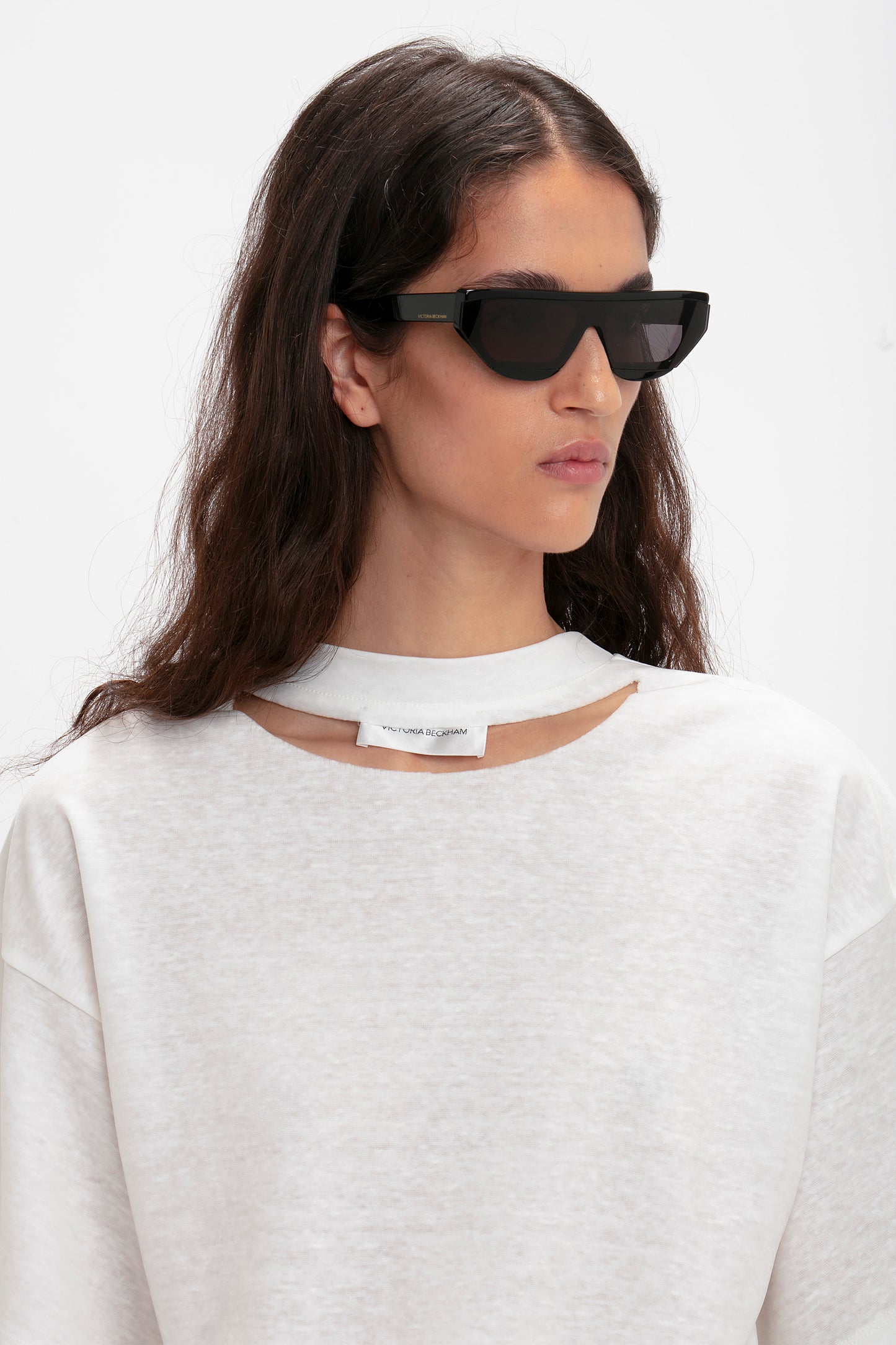 Person with long, dark hair wearing Victoria Beckham Layered Lens Visor Sunglasses In Black and a white sweatshirt with a cut-out neckline, looking to the side against a plain background.