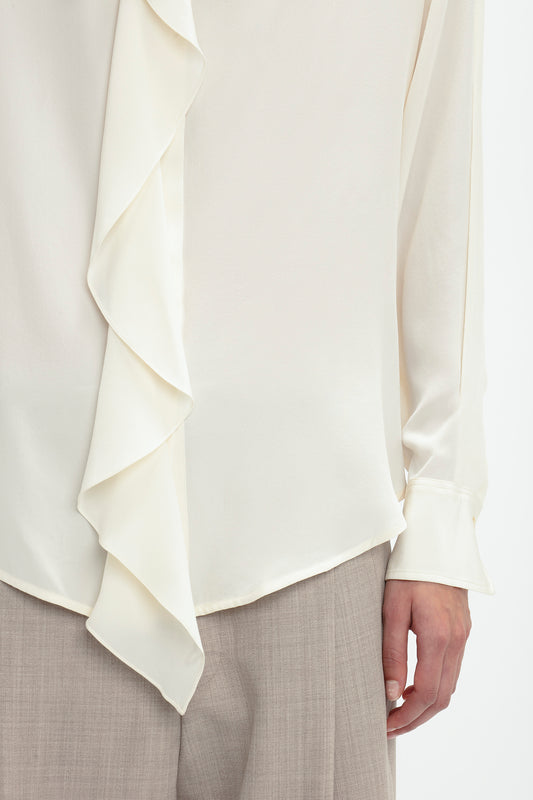 Close-up of a person wearing the Asymmetric Ruffle Blouse In Ivory by Victoria Beckham. The blouse features voluminous sleeves, and is paired elegantly with beige trousers.