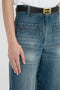 Close-up of a person wearing Victoria Beckham Alina High Waisted Jean In Indigrey Wash, featuring seventies-style pockets and a black belt with a gold buckle, white shirt partially visible.