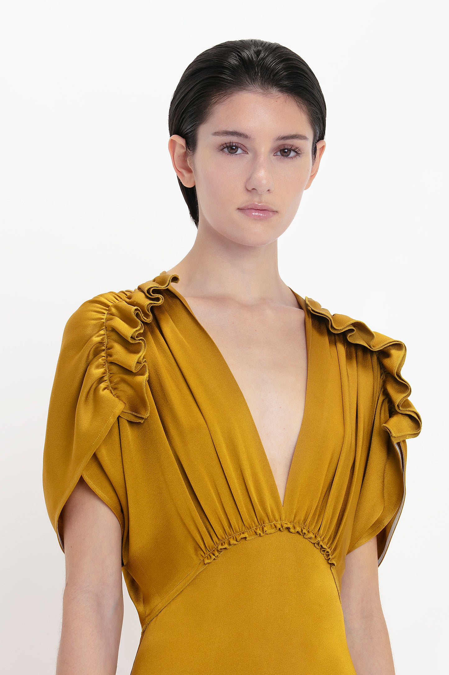 A person with short dark hair is wearing a V-Neck Ruffle Midi Dress In Harvest Gold by Victoria Beckham, looking directly at the camera with a seductive quality. The background is white.