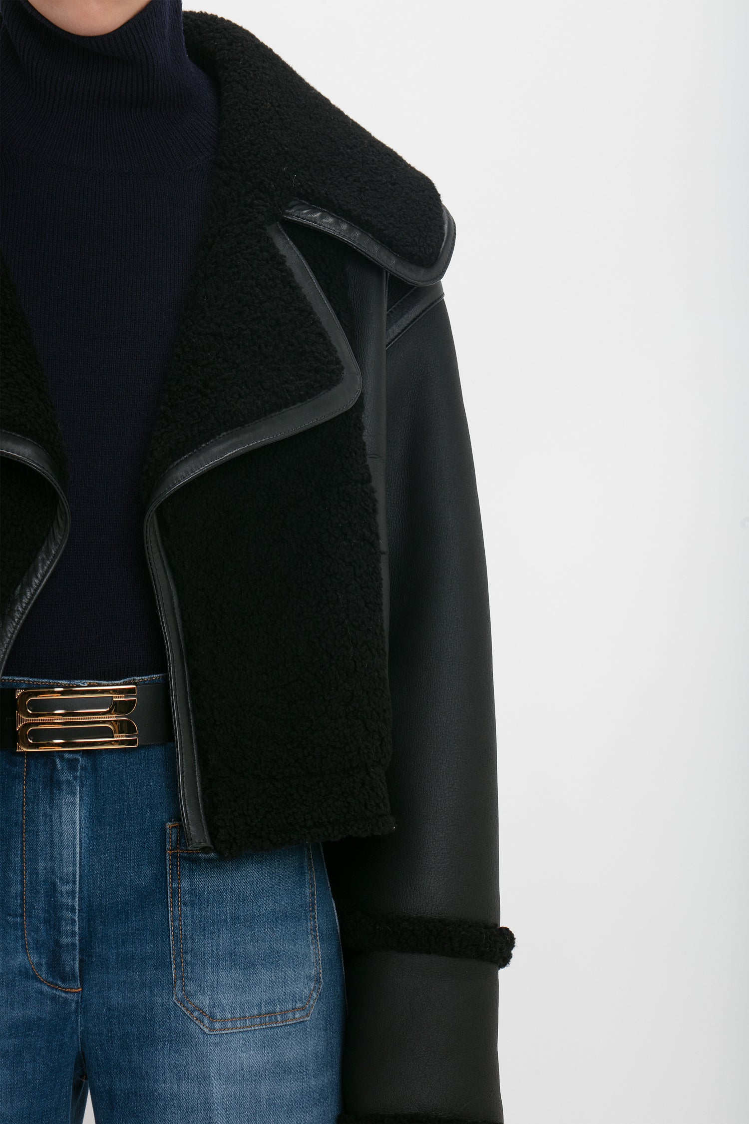 A person wearing a Victoria Beckham Shearling Jacket In Black over a dark turtleneck sweater and blue jeans with a gold-buckled belt.