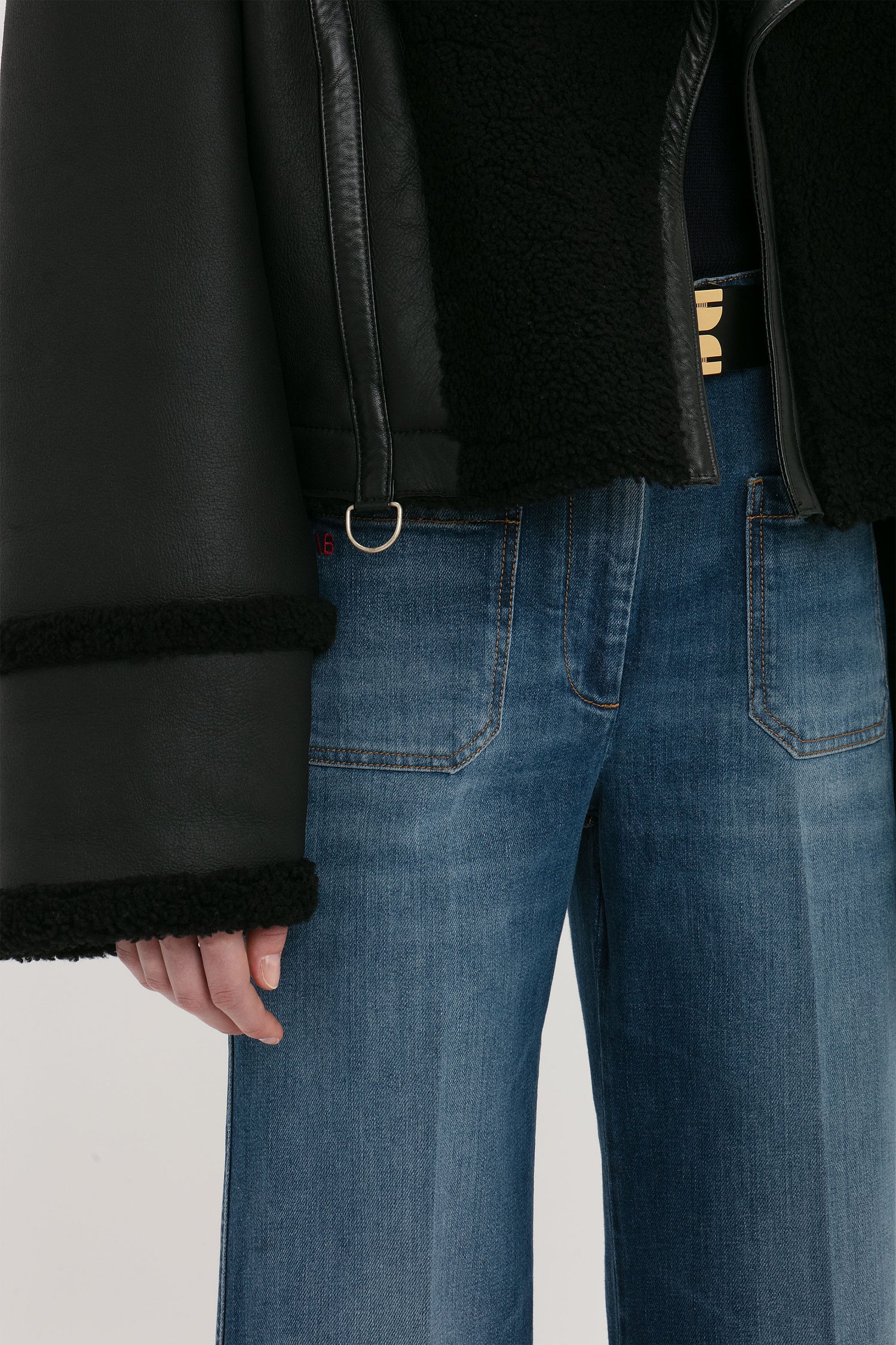 A person wearing blue jeans and a Shearling Jacket In Black by Victoria Beckham with fur trim on the cuffs and edges is shown from mid-thigh to chest.