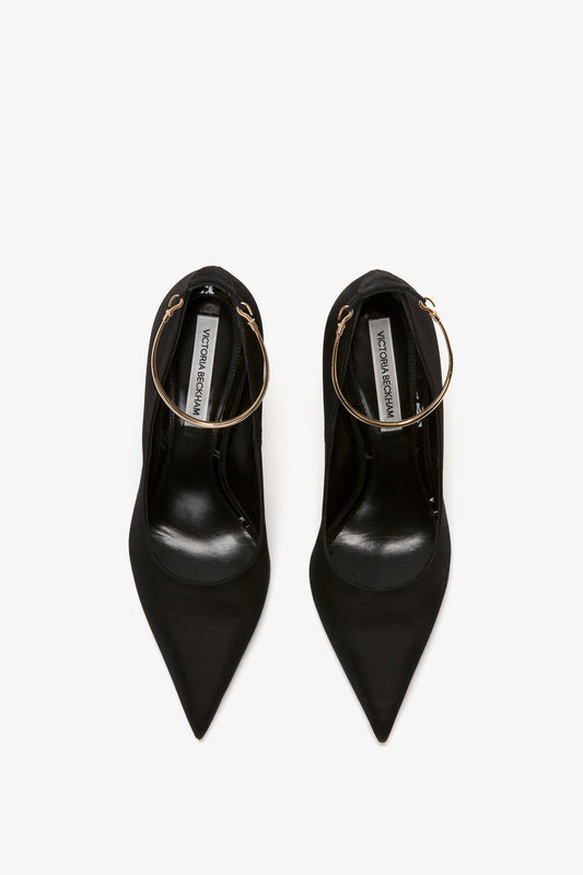 Top view of a pair of Pointy Toe Pump in Black Satin by Victoria Beckham with gold metal ankle bracelets and 100mm heels, placed against a white background. The inner soles display the brand name.