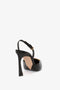 A black Pointy Toe Sling back in Black Calf shoe from Victoria Beckham with a strap around the ankle is shown from the back. The sole of the shoe has a light beige color. The heel is tall and slim with a unique curvature, finished with an elegant chain detail along the strap.