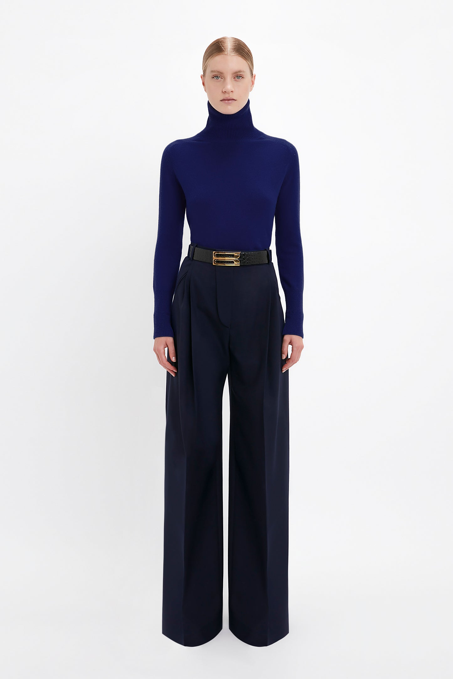 A person stands against a white background wearing a stylish Polo Neck Jumper In Navy by Victoria Beckham and high-waisted, wide-leg navy trousers with a black belt.