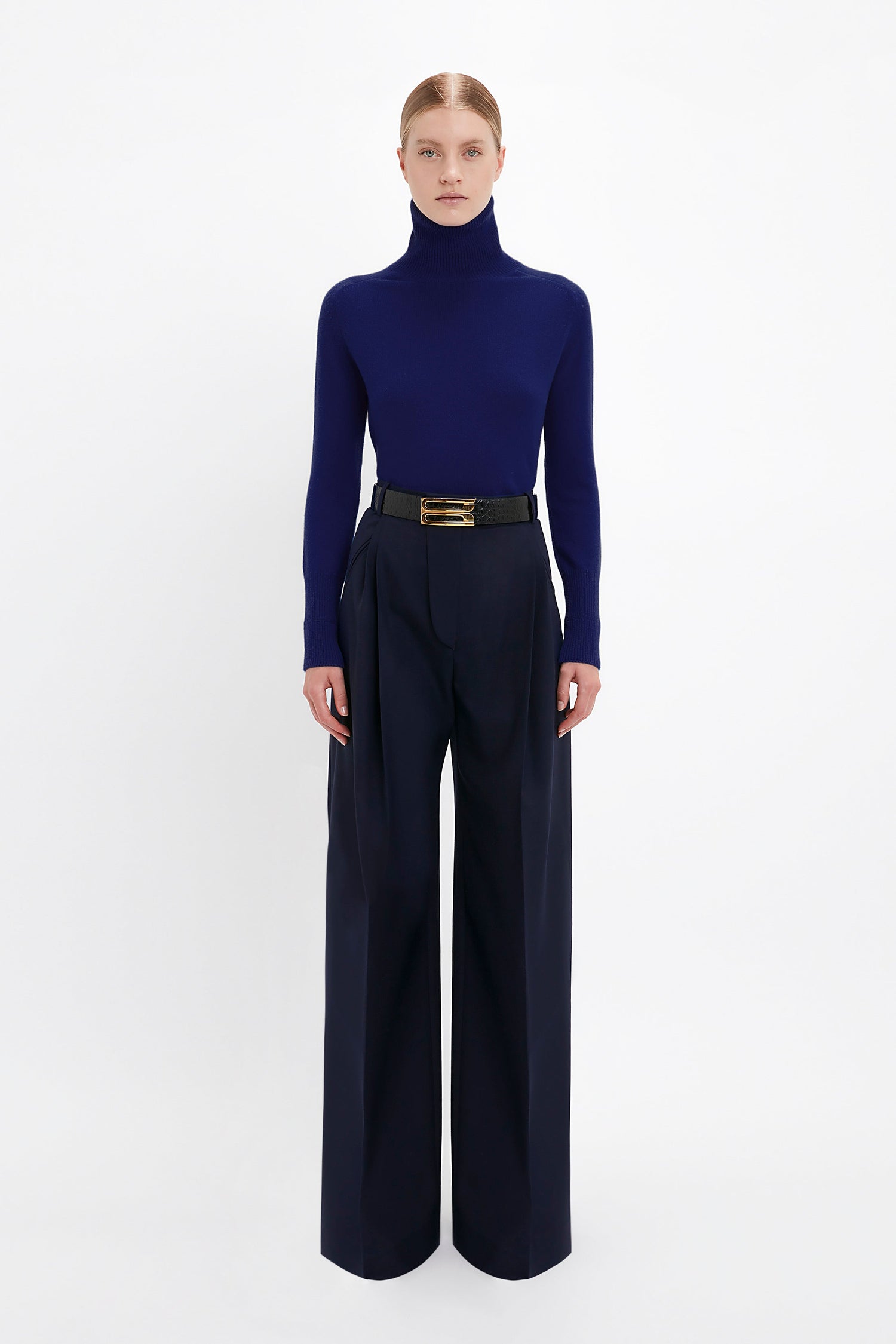 A person stands against a plain white background, wearing a dark blue turtleneck sweater tucked into high-waisted, Wide Leg Trouser In Midnight by Victoria Beckham. The trousers feature pleated front legs and are accessorized with a black belt, creating a sleek midnight look.