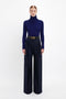 A person stands against a plain white background, wearing a dark blue turtleneck sweater tucked into high-waisted, Wide Leg Trouser In Midnight by Victoria Beckham. The trousers feature pleated front legs and are accessorized with a black belt, creating a sleek midnight look.