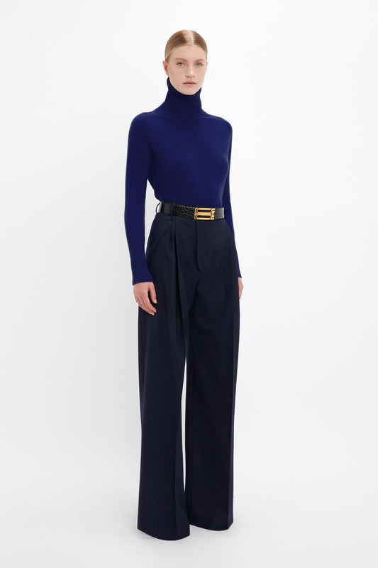 A person is standing against a plain white background wearing a dark blue turtleneck, Victoria Beckham's Wide Leg Trouser In Midnight, and a wide black belt with a gold buckle.