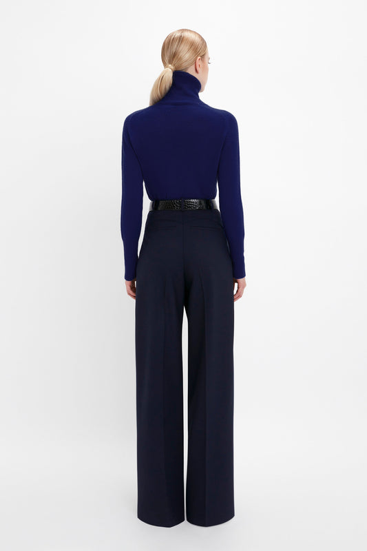 A person stands facing away, wearing a navy blue turtleneck sweater and the Wide Leg Trouser In Midnight by Victoria Beckham, all set against a pristine white background.