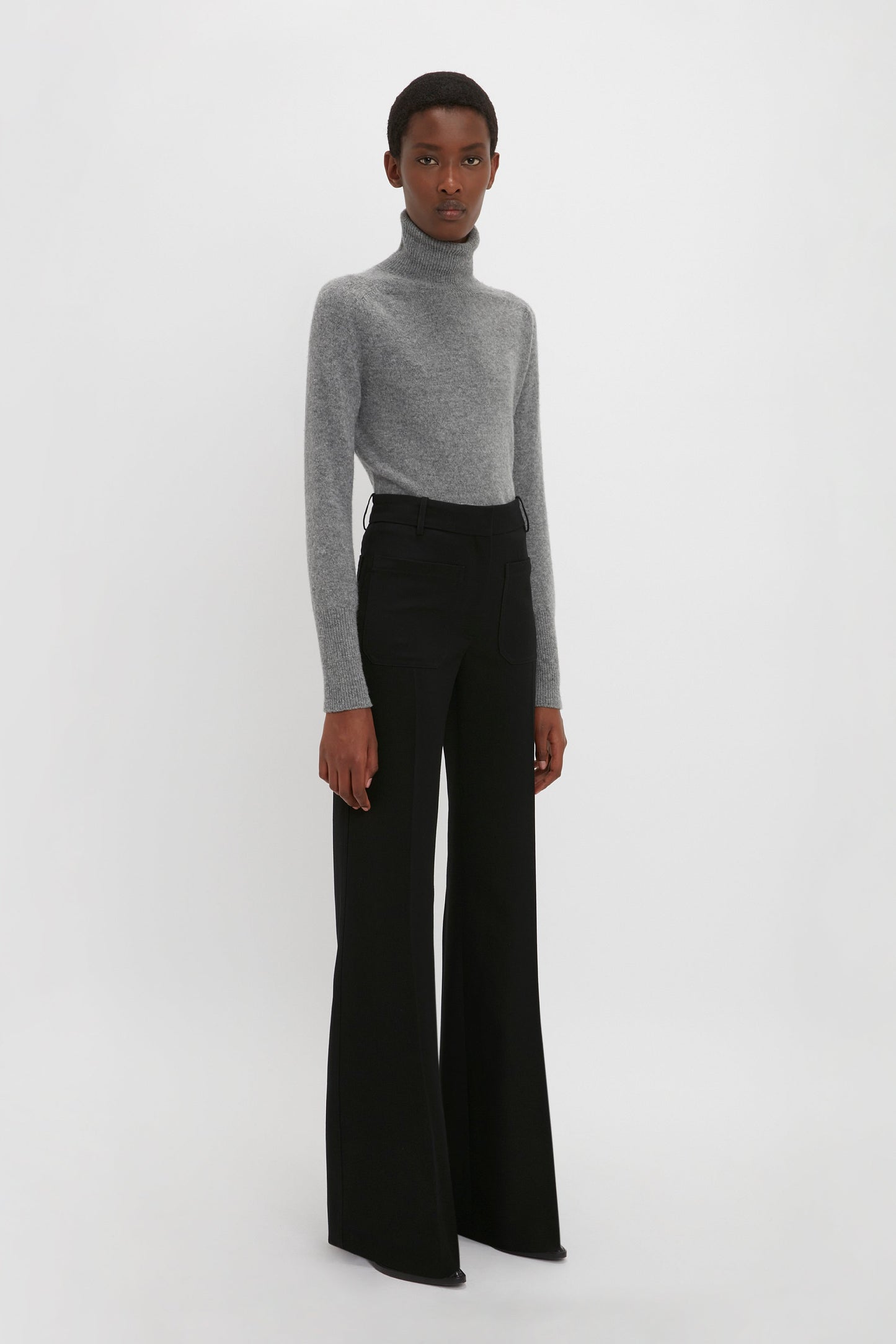 A person in a Victoria Beckham Polo Neck Jumper In Grey Melange and black wide-leg pants stands against a plain white background, showcasing versatile styling.