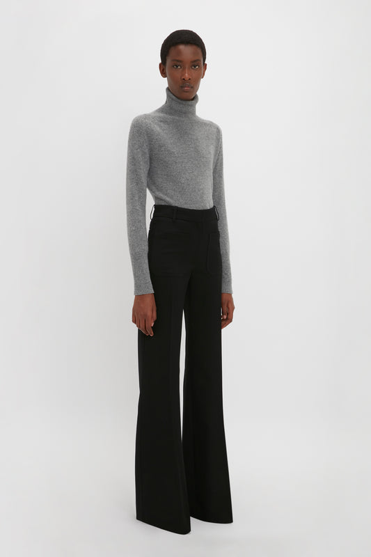 A person stands against a plain background wearing a grey turtleneck sweater and black wide-leg Alina Tailored Trouser In Black, channeling a subtle nod to 70s style reminiscent of Victoria Beckham's chic fashion aesthetic.