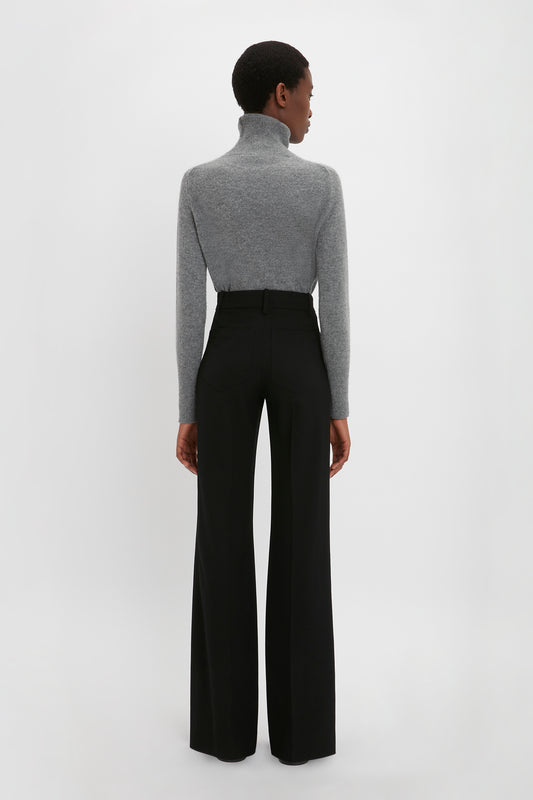 A person with short hair stands facing away, wearing a gray turtleneck sweater and Victoria Beckham's Alina Tailored Trouser in Black, reminiscent of 70s style against a plain white background.