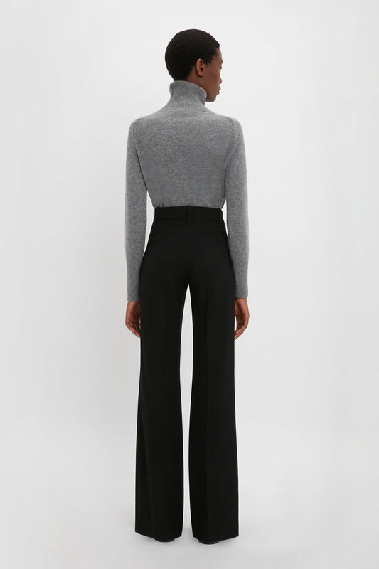 Person facing away from the camera, wearing a versatile Polo Neck Jumper In Grey Melange by Victoria Beckham and high-waisted black wide-leg pants against a plain white background.