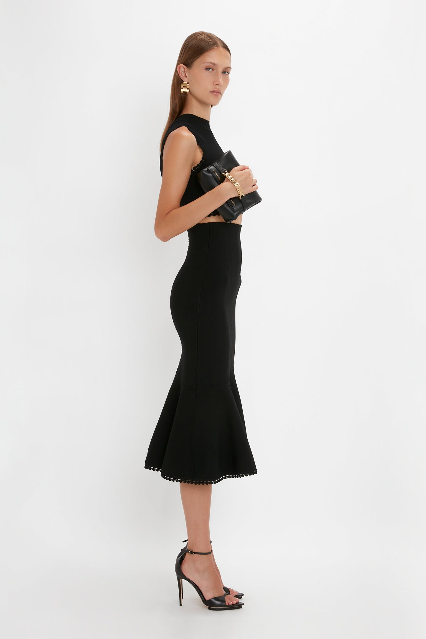 A woman in a sleek, sleeveless black VB Body Scallop Trim Tank Top In Black by Victoria Beckham and high heels stands against a white background, holding a black clutch—a true summer essential.