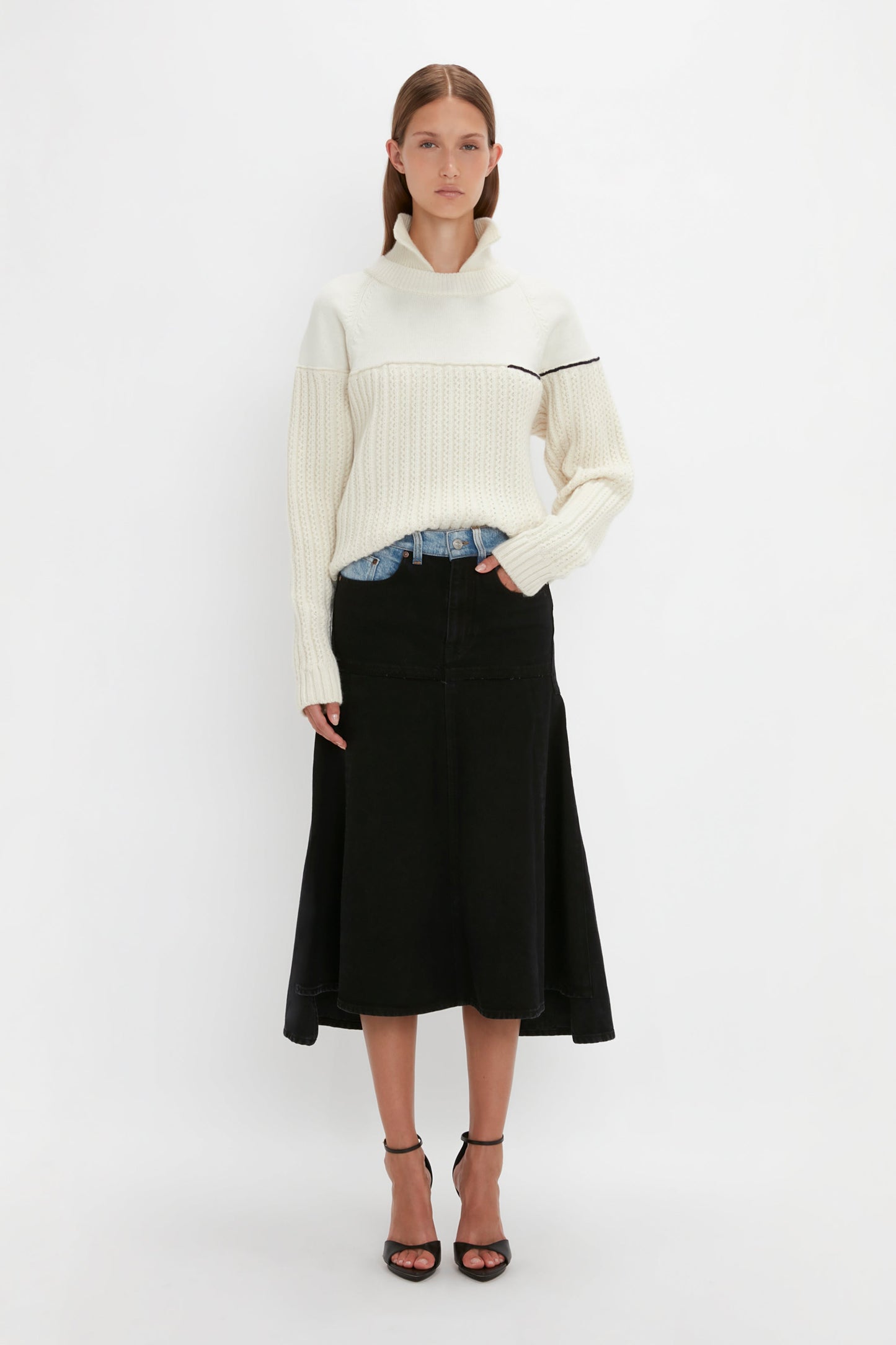 A person stands against a plain white background wearing a Victoria Beckham Collar Detail Jumper In Natural with a black stripe, black skirt, denim jeans, and black heels. One hand is in the pocket of the jeans.