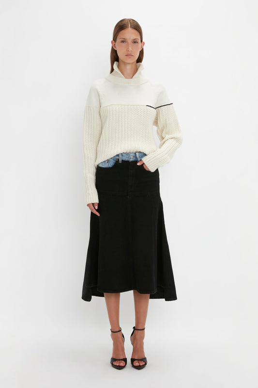 A person stands against a plain white background wearing a Victoria Beckham Collar Detail Jumper In Natural with a black stripe, black skirt, denim jeans, and black heels. One hand is in the pocket of the jeans.