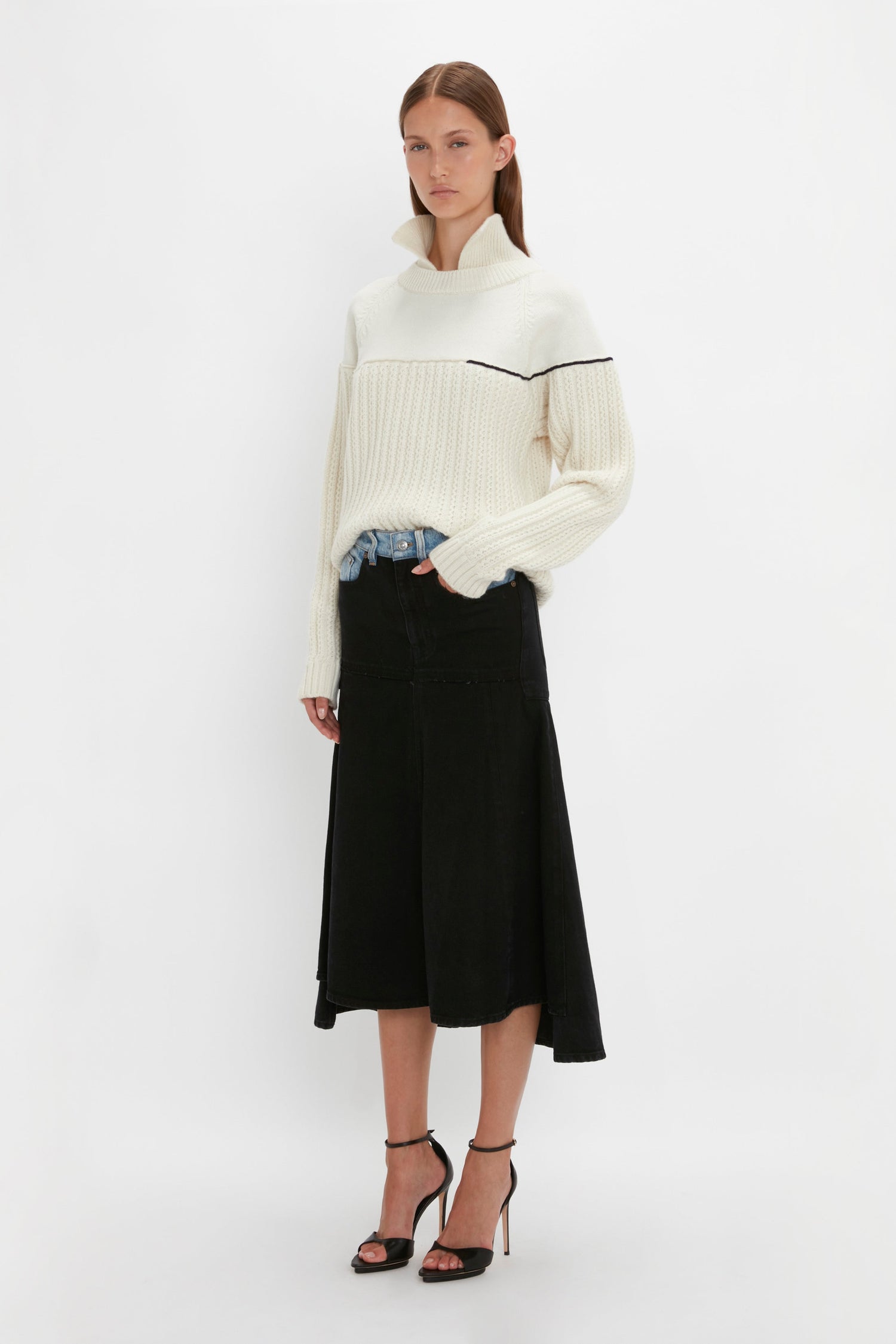 A person stands against a white background wearing a Victoria Beckham Collar Detail Jumper In Natural, a black midi skirt, and black high-heeled sandals.