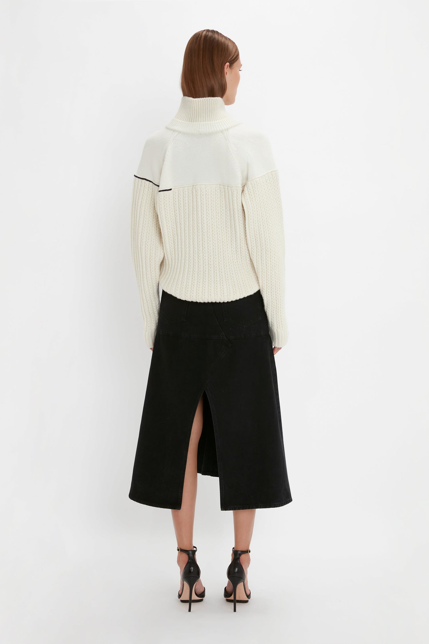 A person with short hair, wearing a Collar Detail Jumper In Natural by Victoria Beckham and a black skirt with a front slit, stands facing away on a white background. They are also wearing black high-heeled sandals.