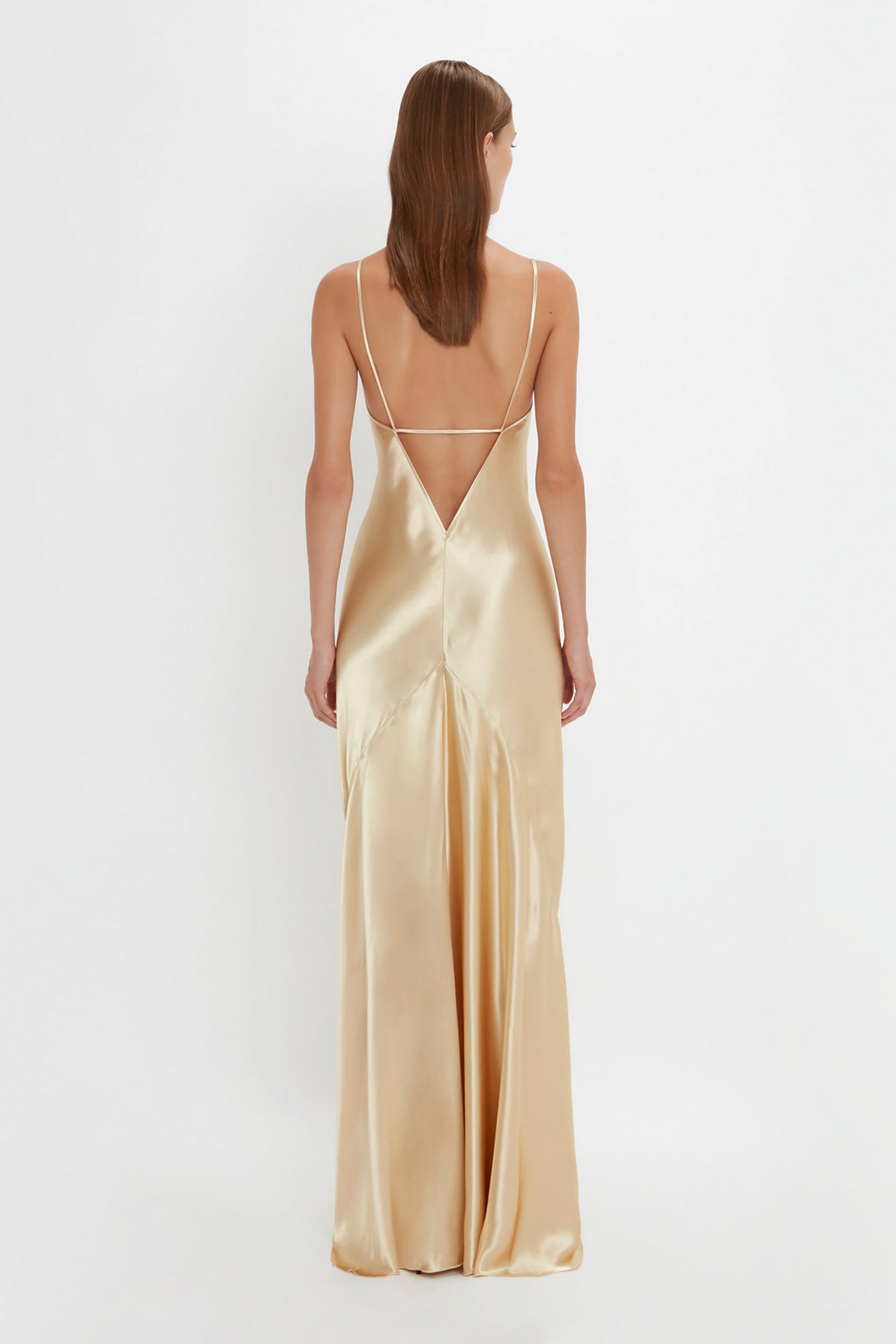 A woman wearing the Victoria Beckham Exclusive Floor-Length Cami Dress In Gold with thin straps, seen from behind against a plain white background, exudes a 1990s-inspired elegance.