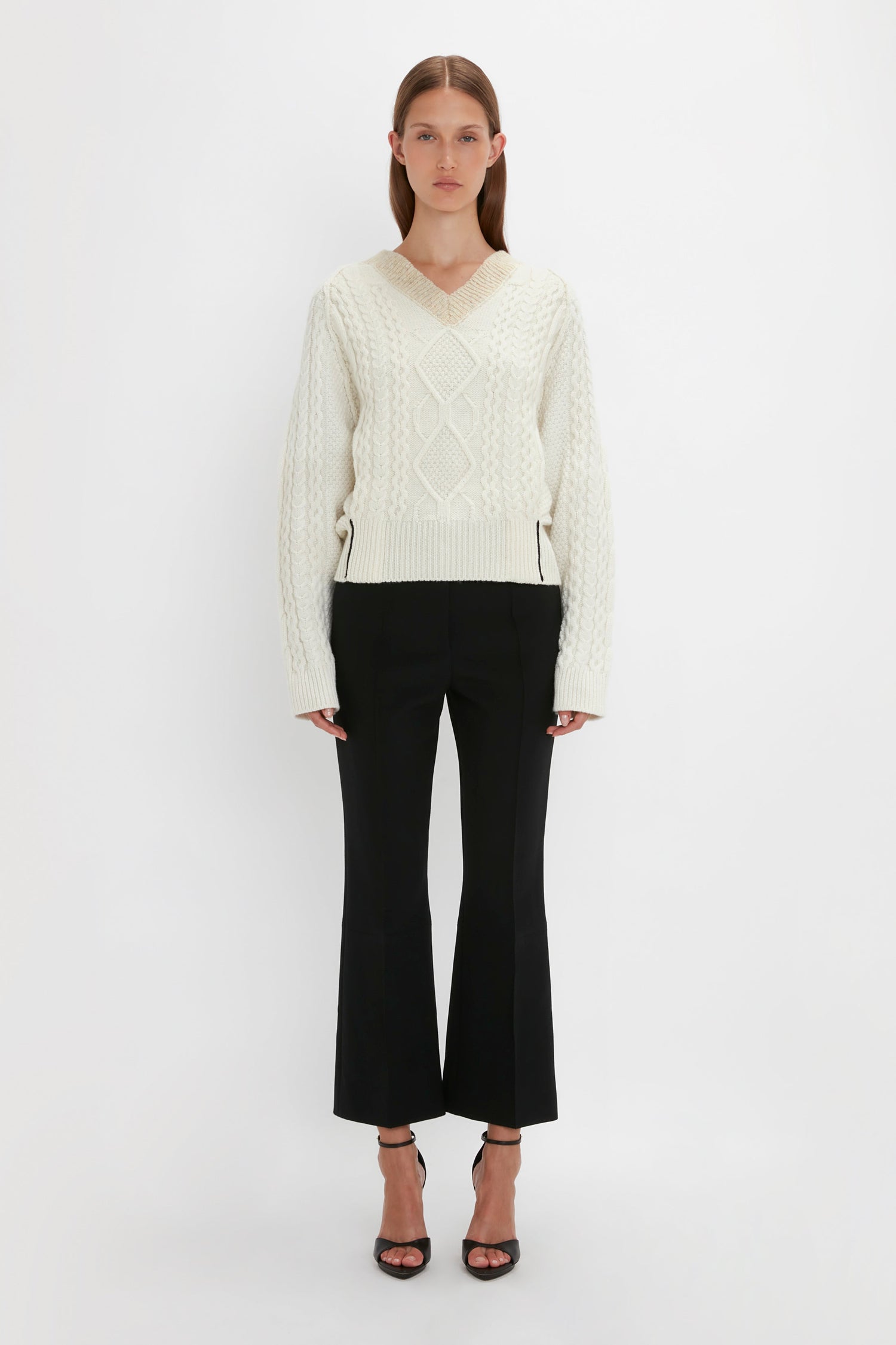 A person stands against a plain white background, wearing a Victoria Beckham V-Neck Jumper In Natural with a diamond pattern and black pants, paired with black open-toe heels.