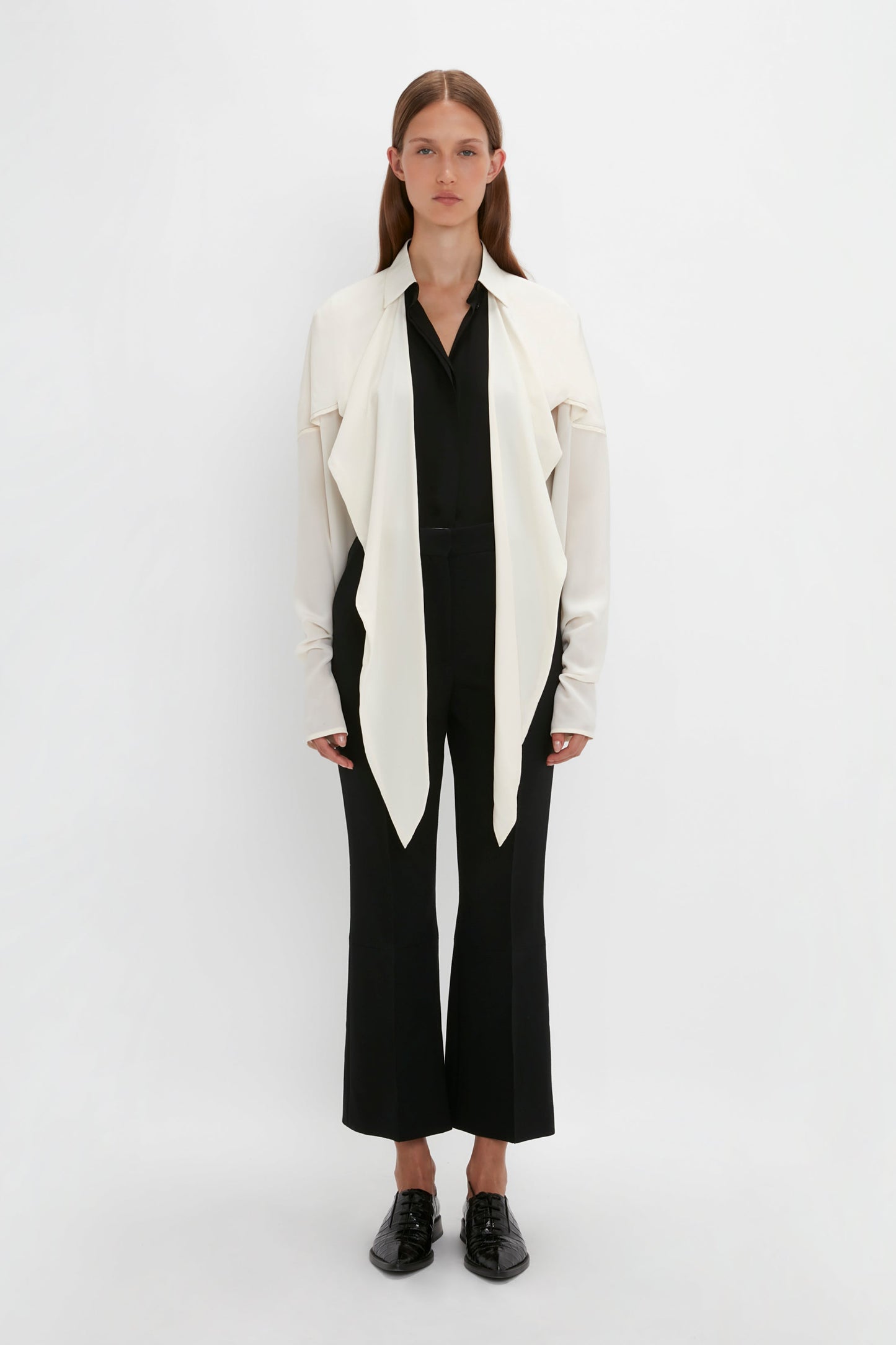 Person standing against a white background, wearing a long-sleeved white jacket with a Victoria Beckham Oversized Bow Detail Blouse In Vanilla over a black shirt and black pants, and black shoes. The dramatic finishing touch adds a feminine flourish to the entire ensemble.