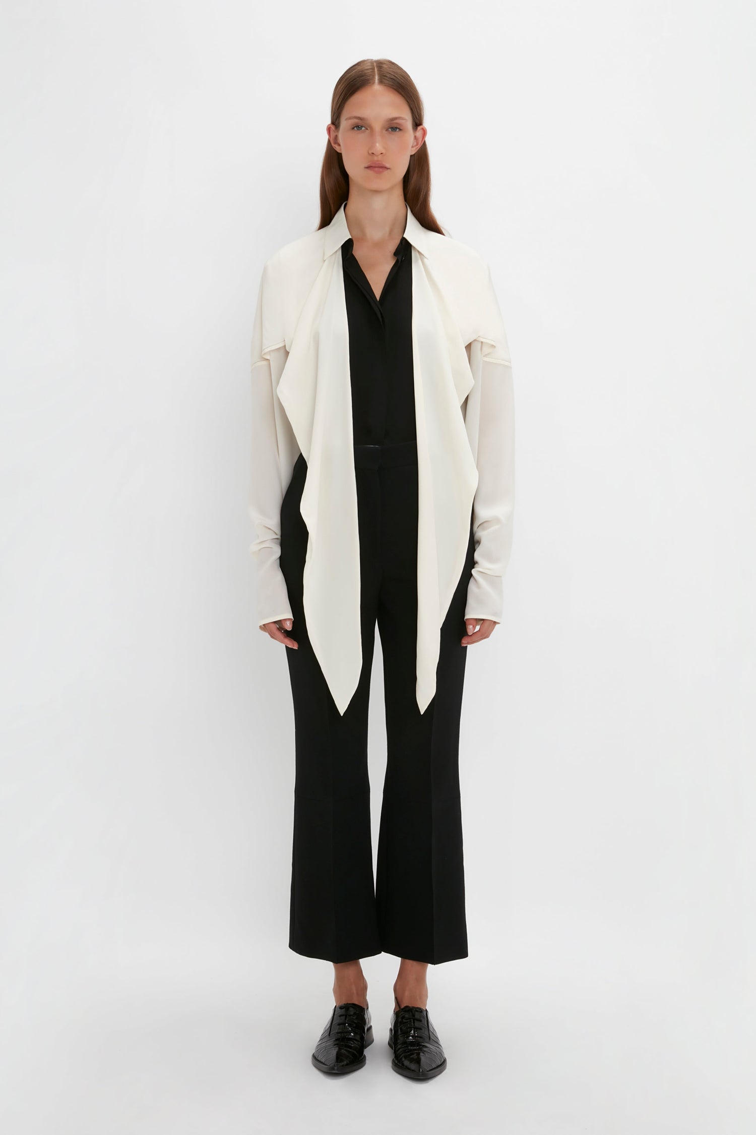 Person standing against a white background, wearing a long-sleeved white jacket with a Victoria Beckham Oversized Bow Detail Blouse In Vanilla over a black shirt and black pants, and black shoes. The dramatic finishing touch adds a feminine flourish to the entire ensemble.