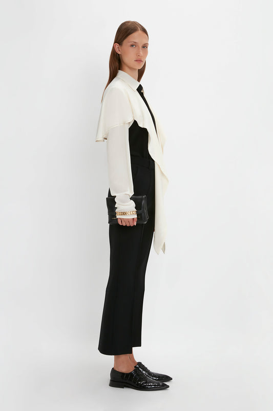 A woman with long hair stares ahead wearing a Victoria Beckham Oversized Bow Detail Blouse In Vanilla and a black and white outfit, holding a black clutch, against a white background. She pairs her look with black shoes and gold bracelets, adding a dramatic finishing touch.