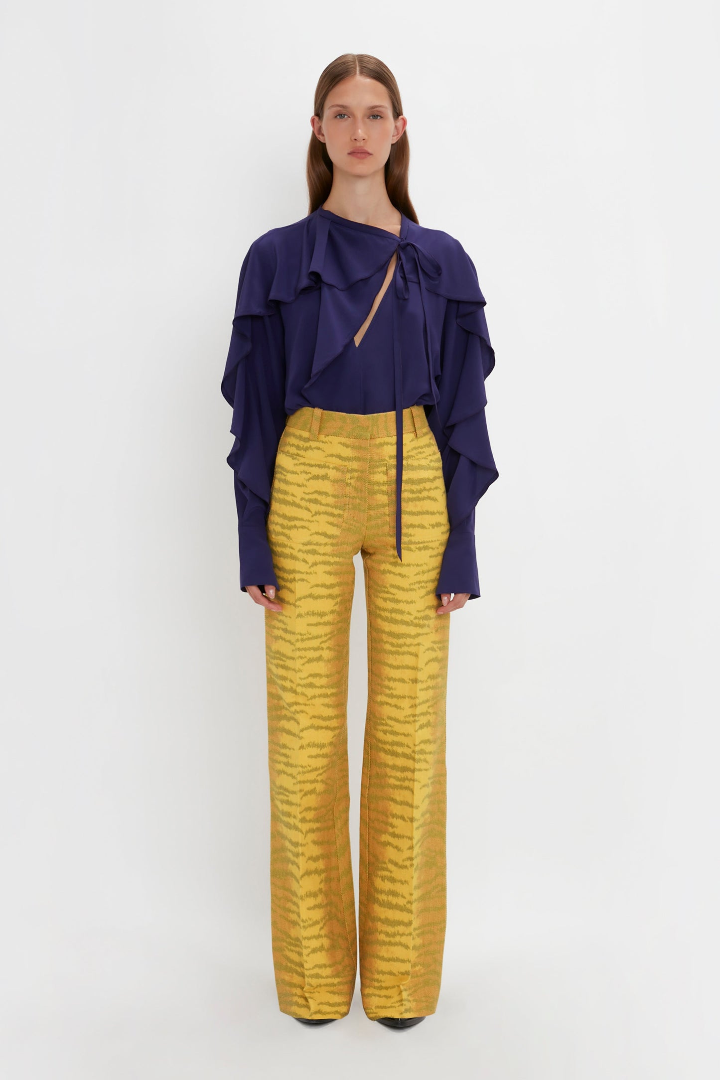 Person poses against a white background wearing the Victoria Beckham Tie Detail Ruffle Blouse In Ultraviolet with a V neckline and yellow high-waisted pants with a striped pattern.