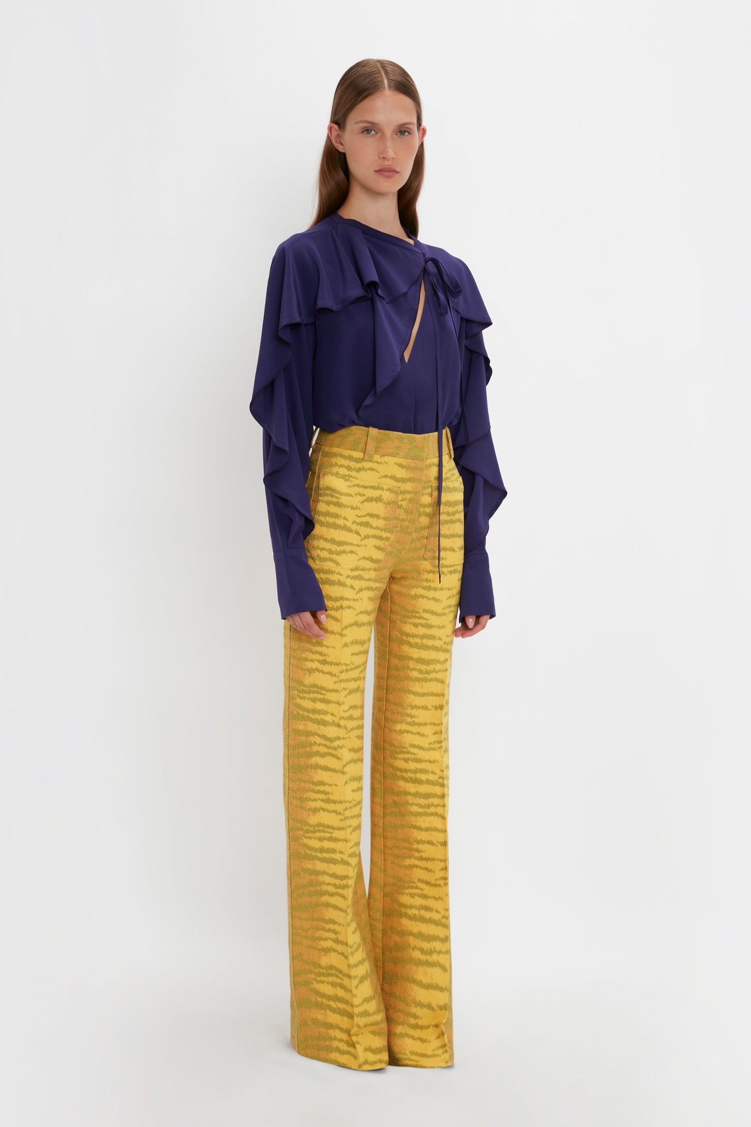 A person stands against a plain background, wearing a Victoria Beckham Tie Detail Ruffle Blouse In Ultraviolet and yellow high-waisted pants with an abstract pattern.