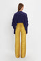 A person is standing with their back to the camera, wearing a Victoria Beckham Tie Detail Ruffle Blouse In Ultraviolet with ruffled sleeves and a V neckline, paired with high-waisted yellow pants featuring a subtle pattern.