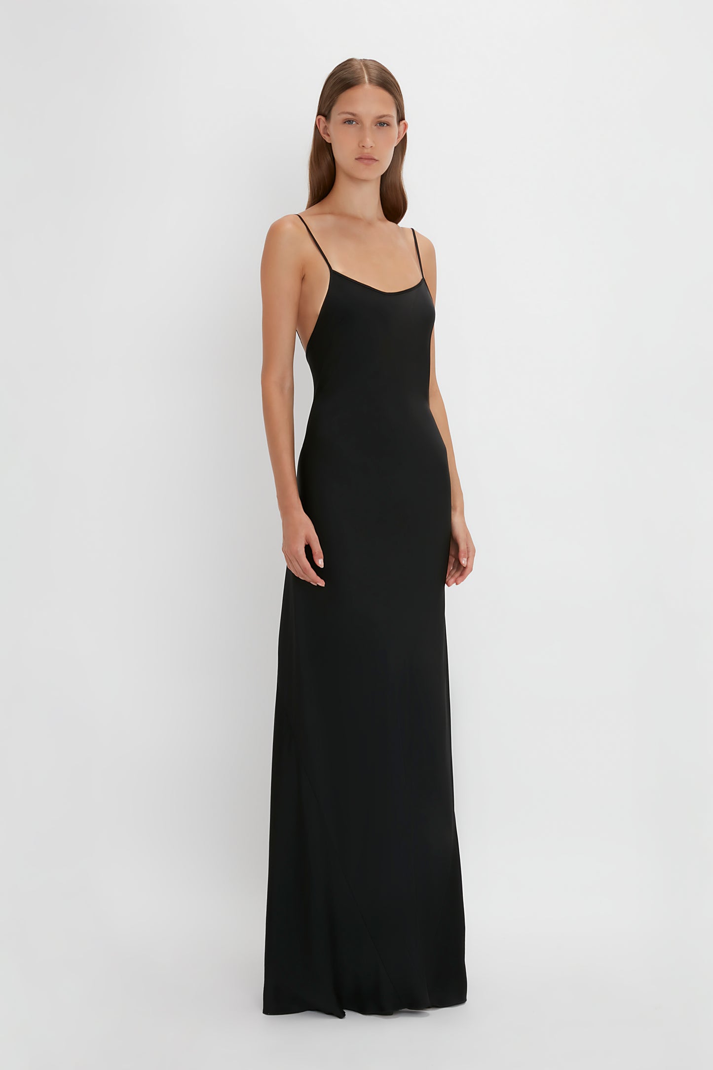 Person standing against a white background, wearing the Victoria Beckham Floor-Length Cami Dress In Black with thin straps.