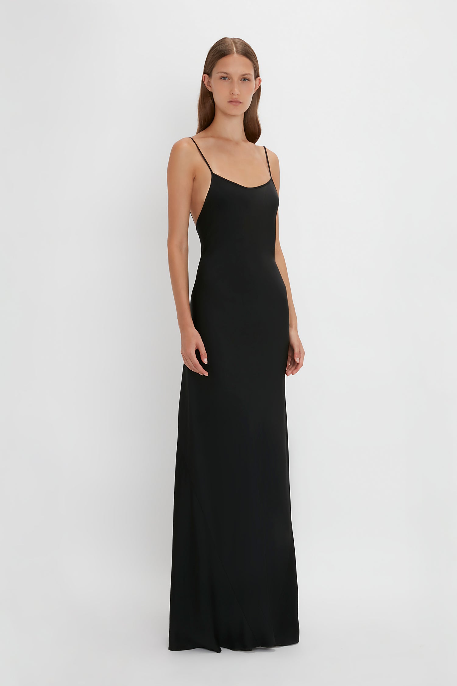 Person standing against a white background, wearing the Victoria Beckham Floor-Length Cami Dress In Black with thin straps.