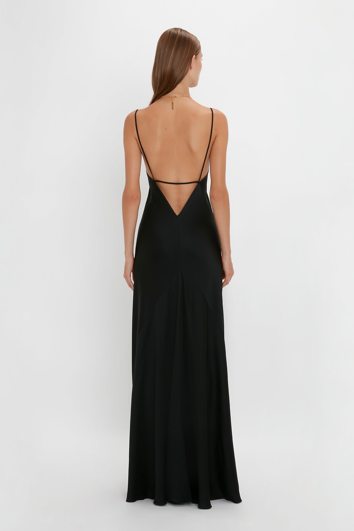 A woman with long brown hair is seen from behind wearing a Floor-Length Cami Dress In Black by Victoria Beckham, standing against a plain white background.