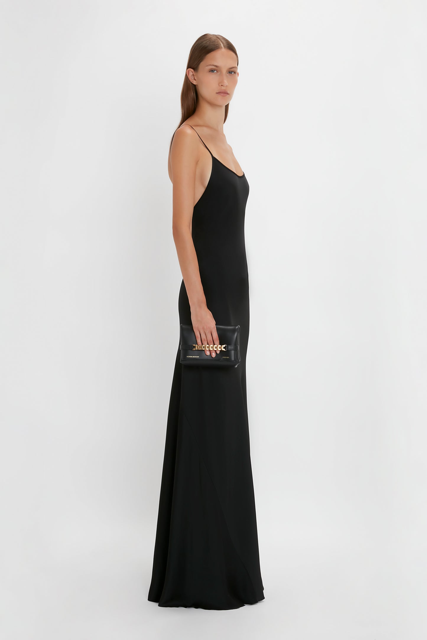 A woman in a shimmering Victoria Beckham Floor-Length Cami Dress In Black stands against a plain white background, holding a black clutch purse.