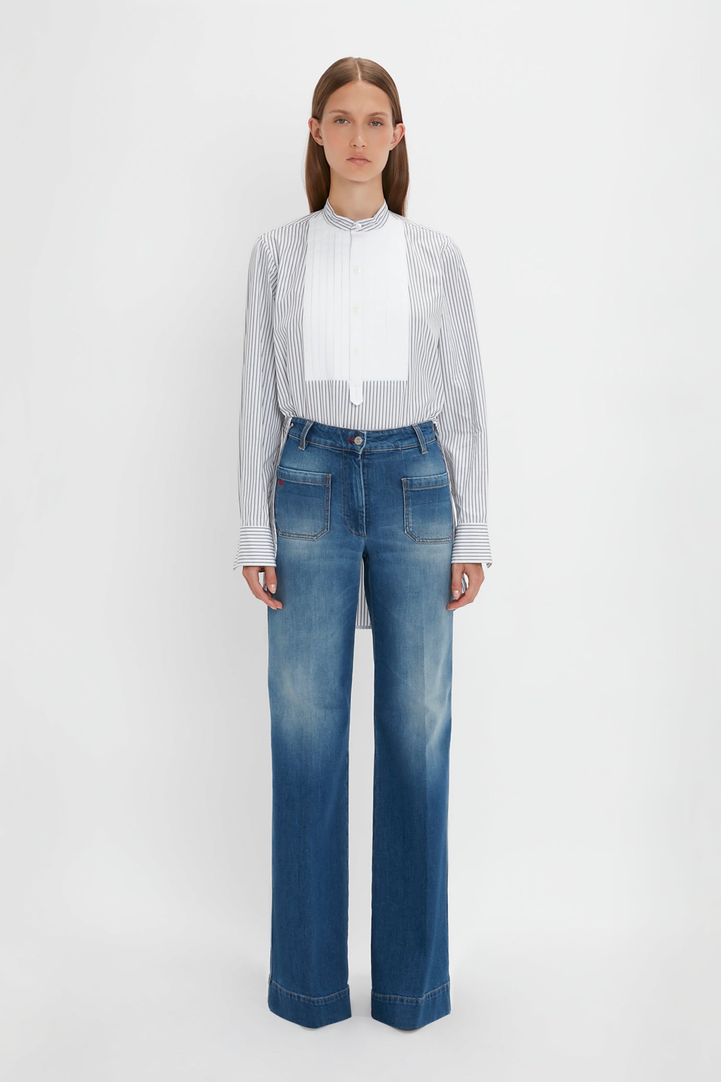 A person stands against a white background, wearing the Victoria Beckham Tuxedo Bib Shirt in Black and Off-White and faded blue wide-leg jeans, embodying relaxed menswear silhouettes.