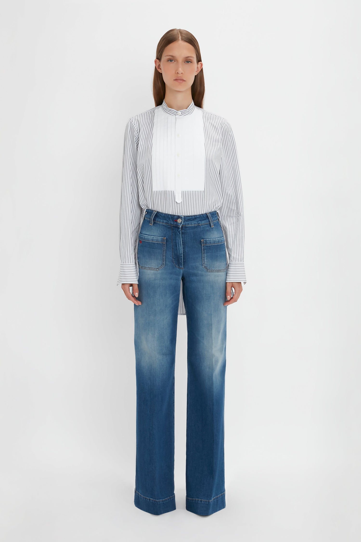 A person stands against a white background, wearing the Victoria Beckham Tuxedo Bib Shirt in Black and Off-White and faded blue wide-leg jeans, embodying relaxed menswear silhouettes.