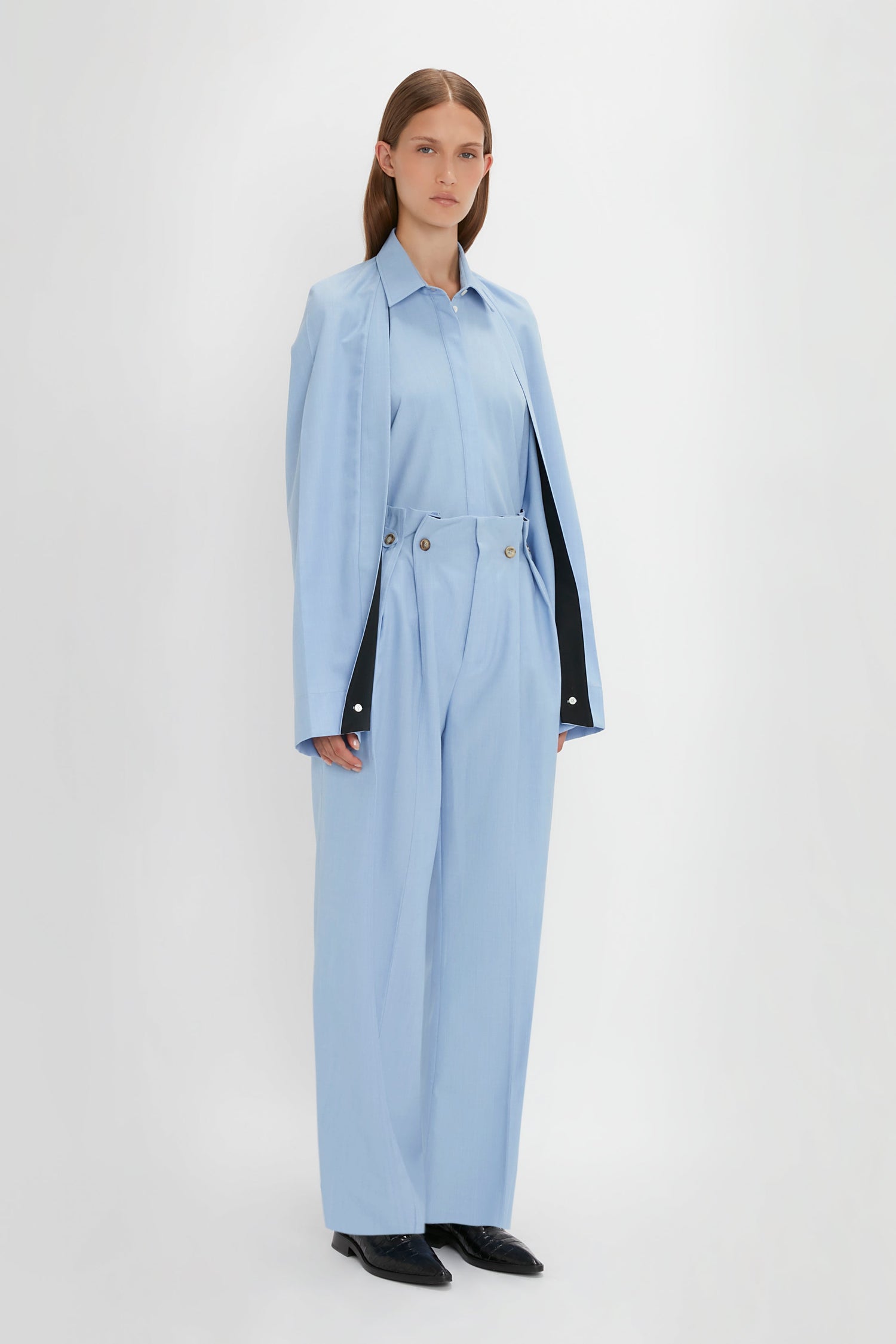 A person is standing against a white background, wearing the Victoria Beckham Pleat Detail Raglan Shirt In Oxford Blue draped over their shoulders and matching high-waisted trousers.