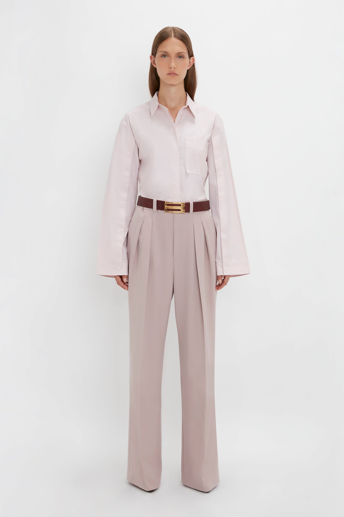 A person with long brown hair stands against a plain white background, wearing a Button Detail Cropped Shirt In Rose Quartz by Victoria Beckham, tan pleated wide-leg trousers, and a brown belt with a gold buckle.