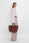 Person wearing a light pink blouse and wide-leg trousers, holding a W11 Medium Tote Bag In Burgundy Leather by Victoria Beckham with adjustable straps, standing against a plain white background.